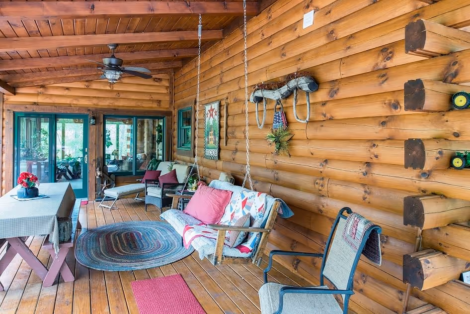 A cozy porch with wooden walls and ceiling, featuring a swing adorned with colorful cushions, a circular rug, and a dining table with a tablecloth. Potted plants and decorative items add warmth to the inviting space.