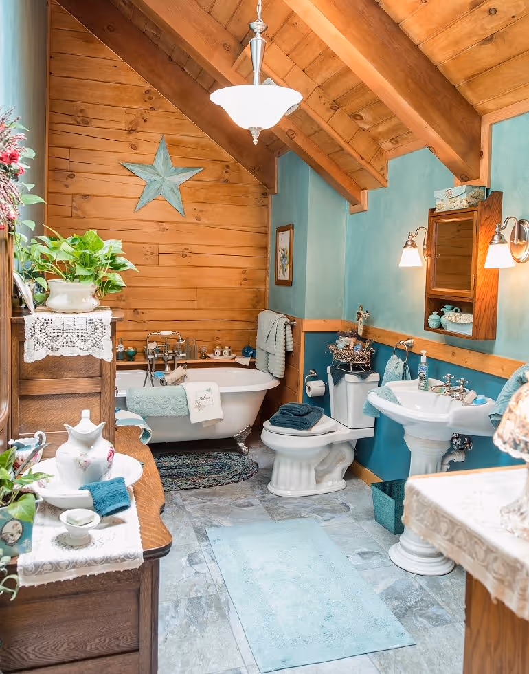 A cozy bathroom featuring wooden accents and teal walls, with a vintage bathtub, white toilet, sink, and decorative plants. The space is illuminated by a pendant light and includes a star decoration on the wall.