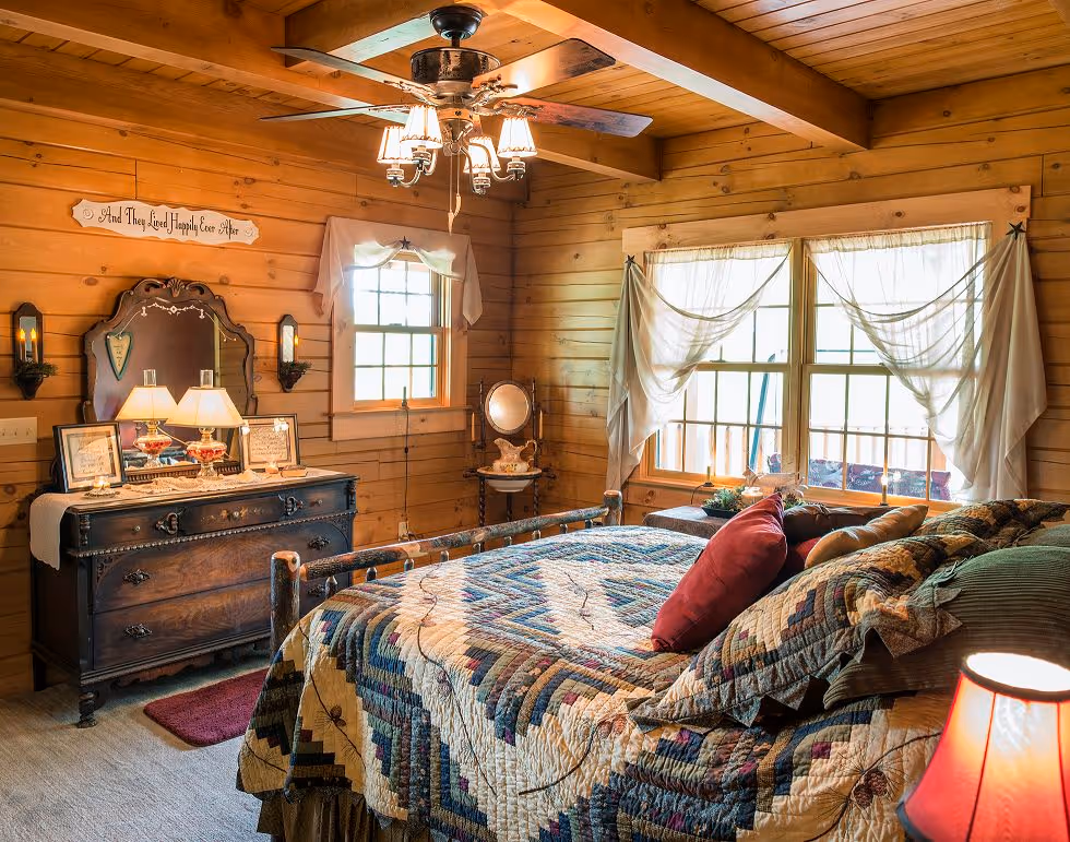 A cozy bedroom with wooden walls, featuring a quilted bedspread in shades of blue and red. A vintage dresser with a lamp and decorative items complements the space, while two windows allow natural light to fill the room.