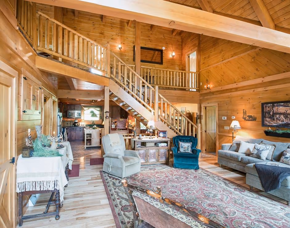 A spacious wooden cabin interior featuring high ceilings, an open staircase, and large windows. The living area includes comfortable seating arrangements, a coffee table, and a rich area rug, creating a cozy atmosphere.