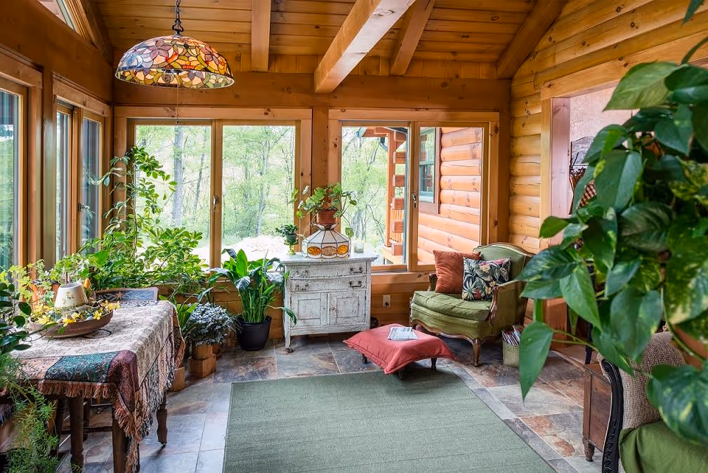 A cozy sunroom featuring wooden beams and large windows, filled with various houseplants. A rustic table with a decorative tablecloth holds potted plants. A green armchair and a red cushion sit on a stone floor, providing a warm atmosphere.