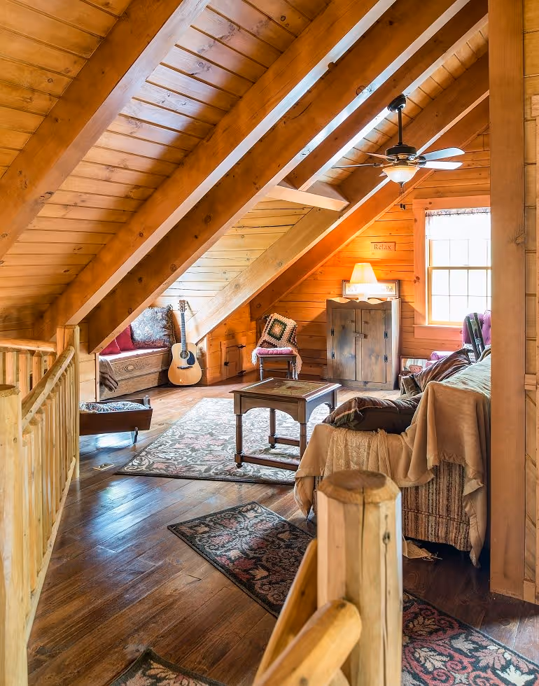 A cozy loft living area featuring wooden beams and flooring, with a guitar on display. A comfortable seating arrangement includes a chair and sofa, complemented by warm lighting and decorative rugs.