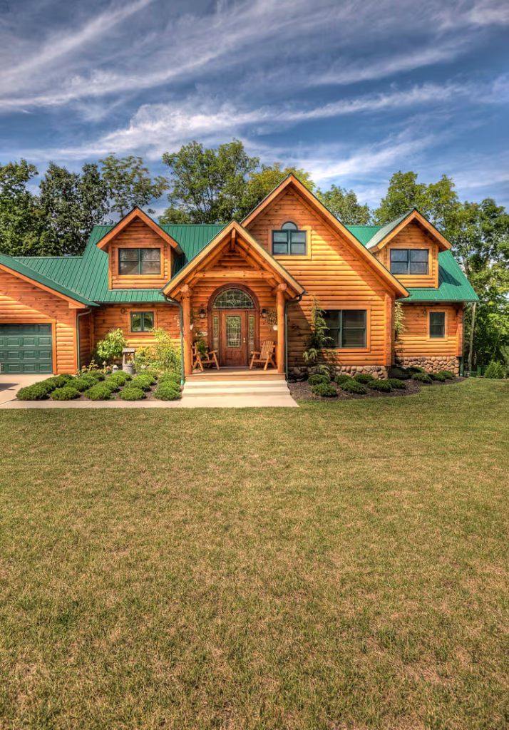 A charming log cabin-style house with a green metal roof, large front porch, and well-maintained landscaping. Surrounding trees add to the serene outdoor setting under a clear blue sky.