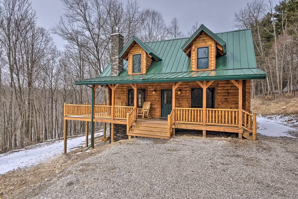 A rustic wooden cabin with a green metal roof surrounded by bare trees. The cabin features a front porch with wooden railings and a stone chimney. A gravel path leads to the entrance, with a light dusting of snow on the ground.