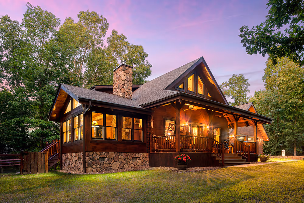A charming wooden house with a stone exterior and multiple large windows, surrounded by lush green trees at dusk, casting a warm glow from the lights inside. A wooden deck with steps leads to the entrance.