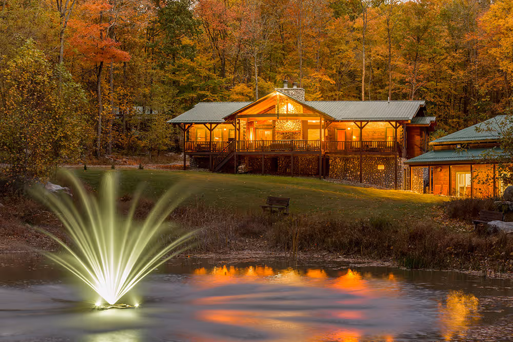 A cozy wooden cabin with large windows is illuminated at dusk, surrounded by vibrant autumn foliage. A colorful fountain shoots water into a tranquil pond in the foreground, reflecting the warm lights of the cabin.