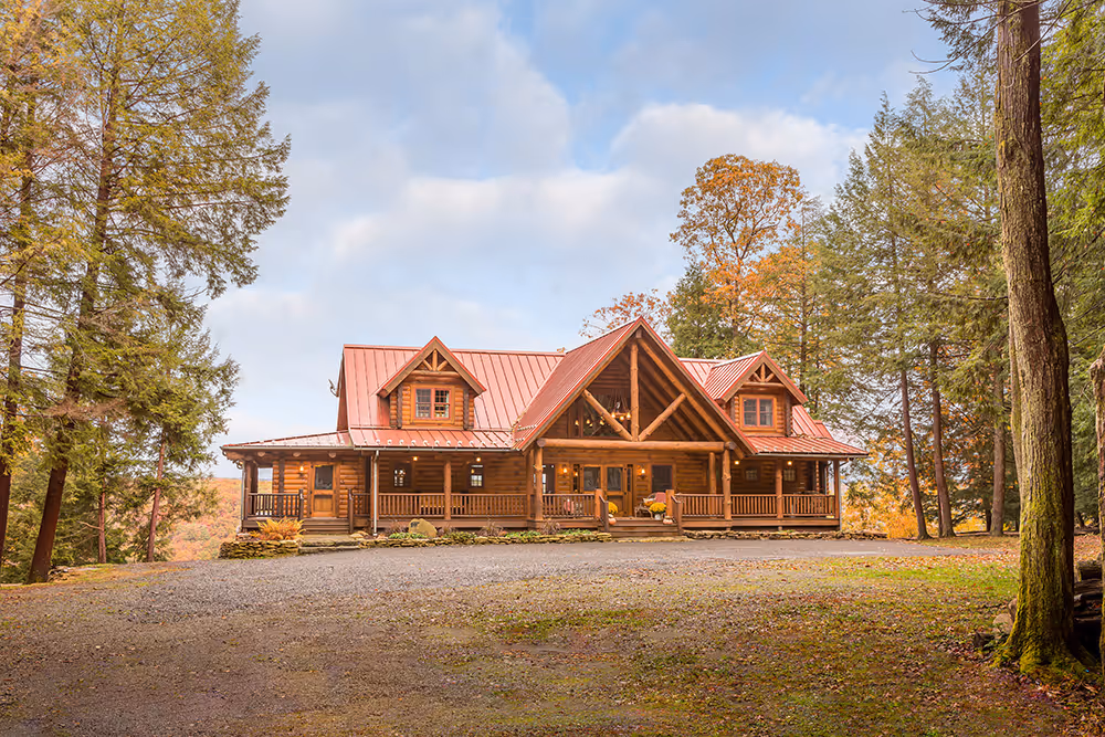 A spacious log cabin with a red metal roof, surrounded by tall pine trees and fall foliage, set on a gravel driveway in a tranquil wooded area. The cabin features a large front porch and multiple gables.