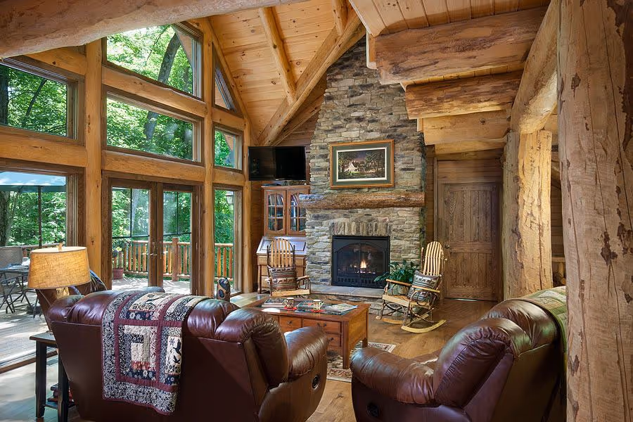 A cozy cabin living room featuring large windows that let in natural light, a stone fireplace, and wooden beams. Two brown leather armchairs face a wooden coffee table, while a quilt drapes over one chair. A rustic ambiance surrounded by trees is evident.
