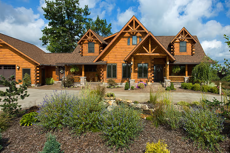 A beautiful log cabin-style home surrounded by lush green landscaping and colorful flowers, with a spacious driveway leading to a warm wooden front porch, set against a backdrop of partly cloudy skies.