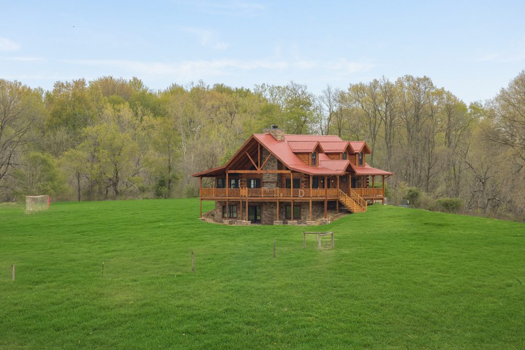 A log home with a red metal roof and stone foundation sitting on a wide, mown green field, with a covered porch across the front and bare early-spring woods behind it.