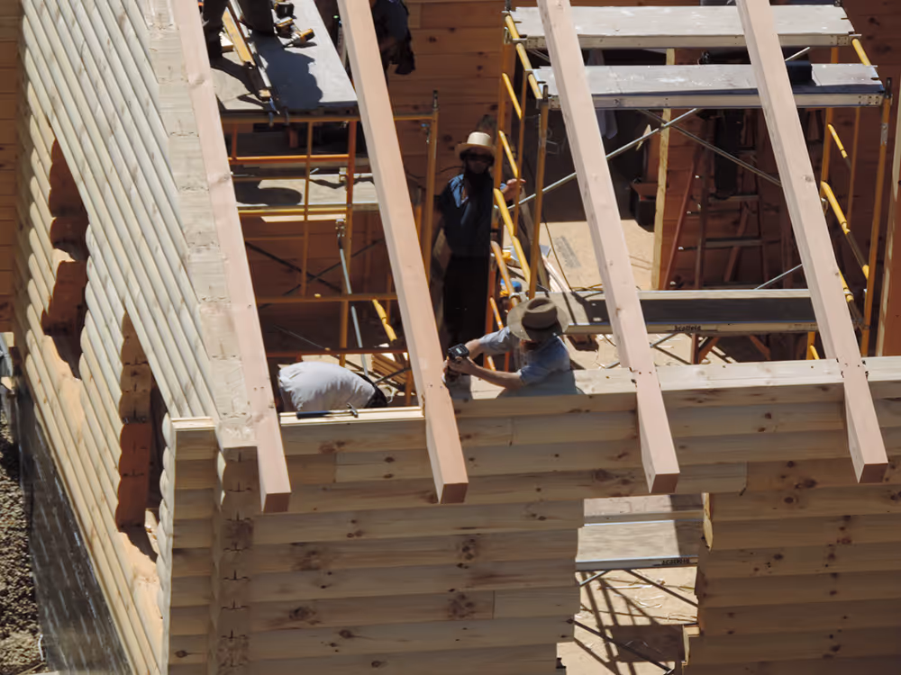 Construction workers installing roof beams on a wooden structure, with scaffolding visible around the building. The scene captures several individuals focused on their tasks under bright sunlight.
