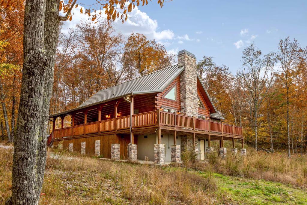 A charming log cabin with a sloped roof, featuring a stone chimney and large wooden porch, surrounded by autumn foliage. The warm tones of the leaves contrast with the rustic wood of the cabin, creating a cozy atmosphere in the serene landscape.