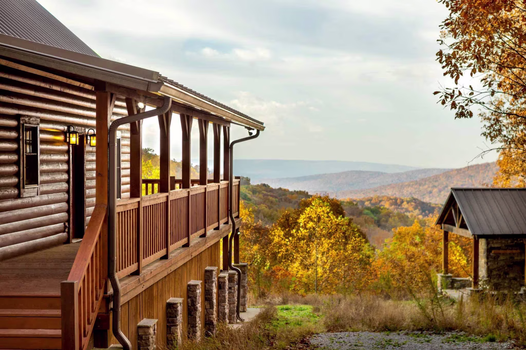 A wooden cabin with a spacious porch overlooks a valley filled with vibrant autumn foliage. The landscape features rolling hills under a partly cloudy sky, offering a peaceful rural retreat.