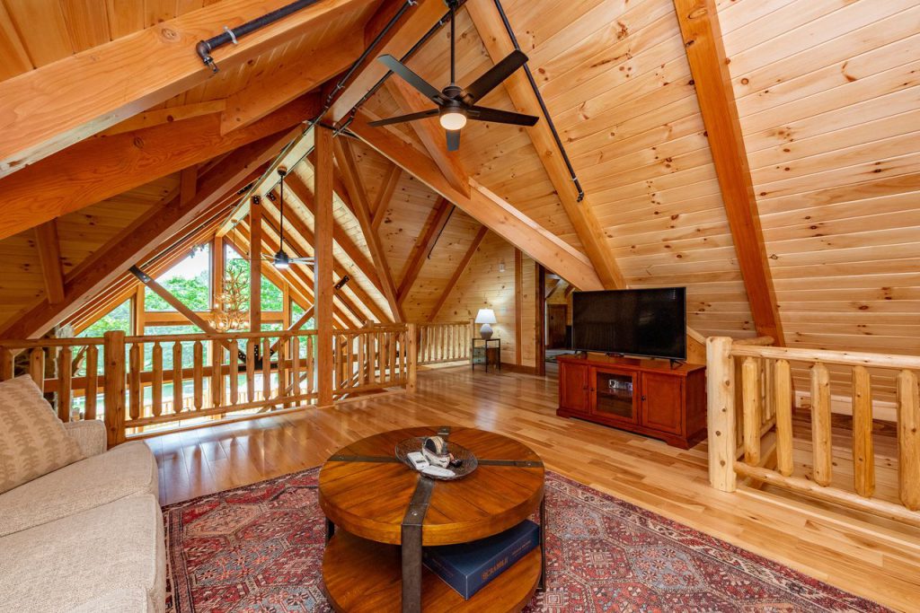 An inviting loft space featuring wooden walls and ceilings, with exposed beams. A cozy seating area includes a light-colored sofa and a round coffee table. A TV is placed in a wooden cabinet, with large windows.