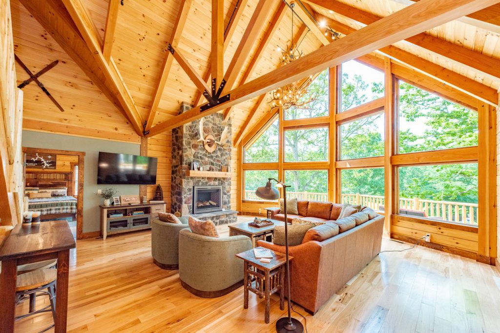 A cozy cabin living room with wooden ceilings and large windows, featuring a stone fireplace and plush seating. Natural light floods the space, highlighting the warm wood tones and inviting ambiance.