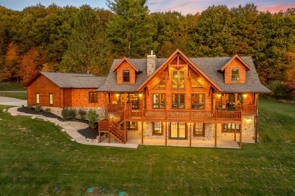 A large, charming log cabin home with multiple gabled roofs, illuminated at dusk. The property features a spacious deck, surrounded by lush green grass and colorful fall trees in the background, creating a picturesque setting.