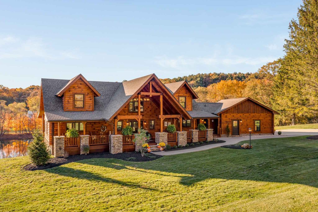 A beautifully crafted log cabin house with a large front porch, surrounded by vibrant autumn trees and a grassy lawn. The two-story home features stone accents and large windows, reflecting a serene rural landscape with a clear blue sky.