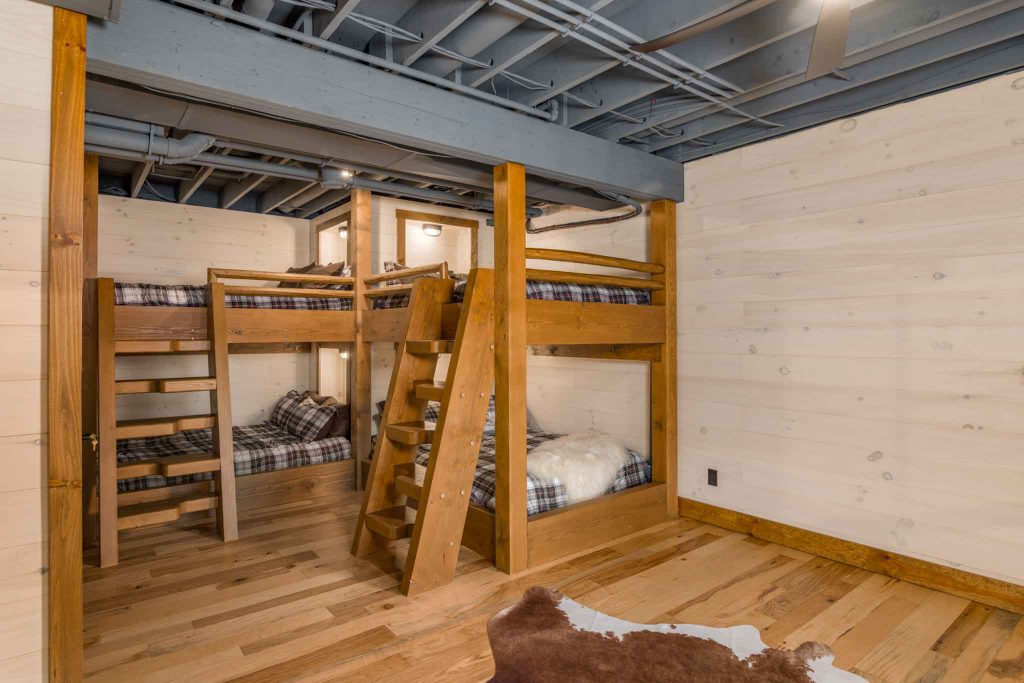 A cozy bedroom featuring two wooden bunk beds with plaid bedding, placed against a light wooden wall. Natural wood flooring and exposed beams add warmth to the serene space, creating an inviting atmosphere for relaxation.