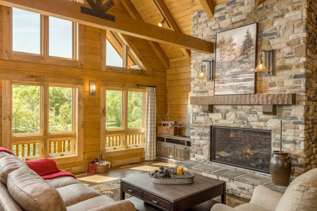 A cozy living room with a high wooden ceiling, featuring large windows overlooking greenery. A stone fireplace with a wooden mantel and a landscape painting above it complements comfortable seating arranged around a dark wooden coffee table.