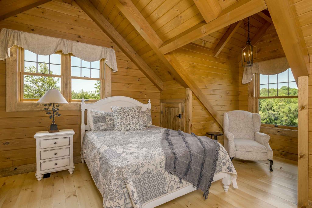 A cozy wooden bedroom featuring a white bed with a decorative gray blanket, a nightstand with a lamp, and a comfortable armchair. Large windows offer a view of greenery outside, enhancing the warm atmosphere.