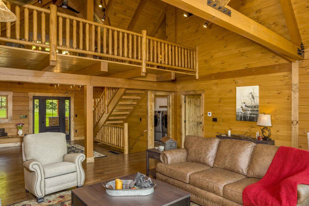 A spacious wooden interior of a log cabin featuring a cozy living area with a brown leather sofa, an armchair, and a coffee table. A staircase leads to a loft area, and large windows allow natural light to fill the space.