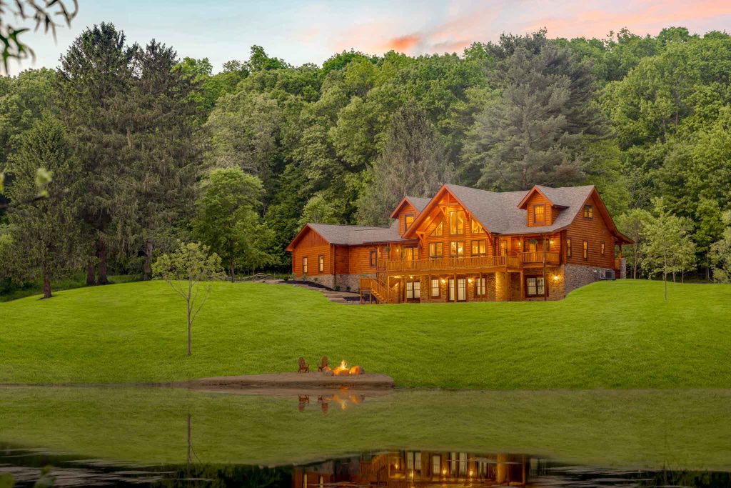 A charming wooden cabin with large windows, nestled on a grassy slope next to a serene lake. Surrounding trees reflect in the water, creating a peaceful, natural setting, with a small fire pit visible in the foreground.