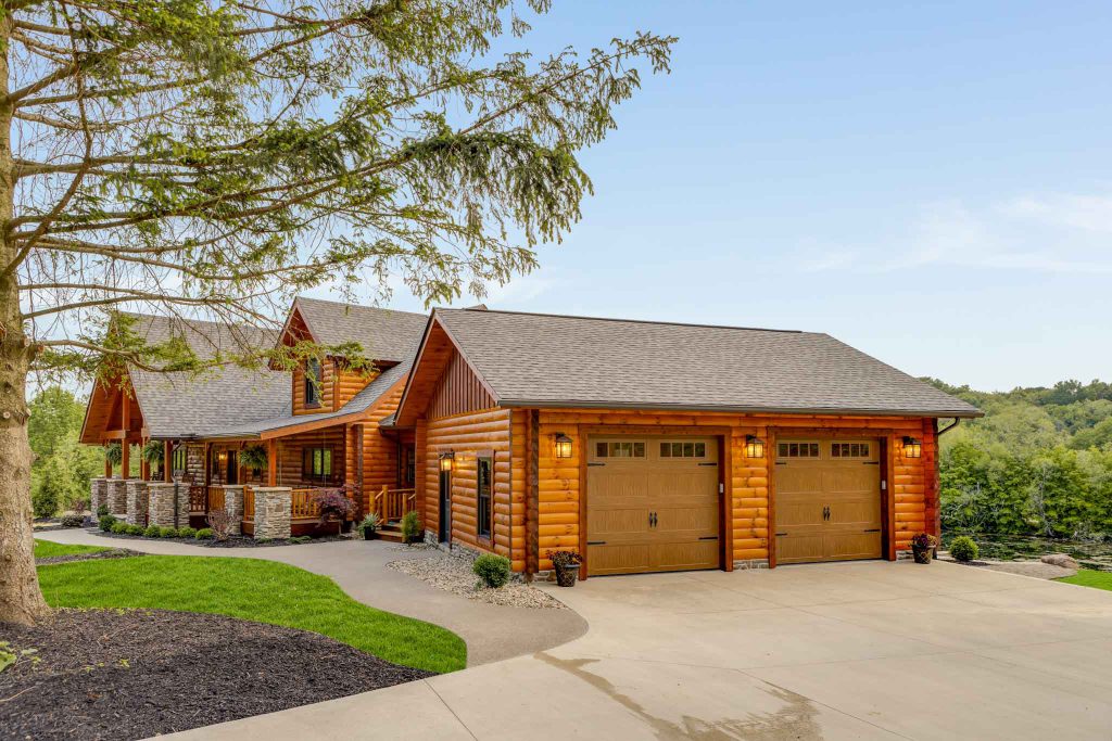 A picturesque log cabin home with a comfortable front porch, set among lush greenery. The house features a gabled roof and two garage doors, with a neatly paved driveway leading up to it. Bright blue sky in the background adds to the serene ambiance.