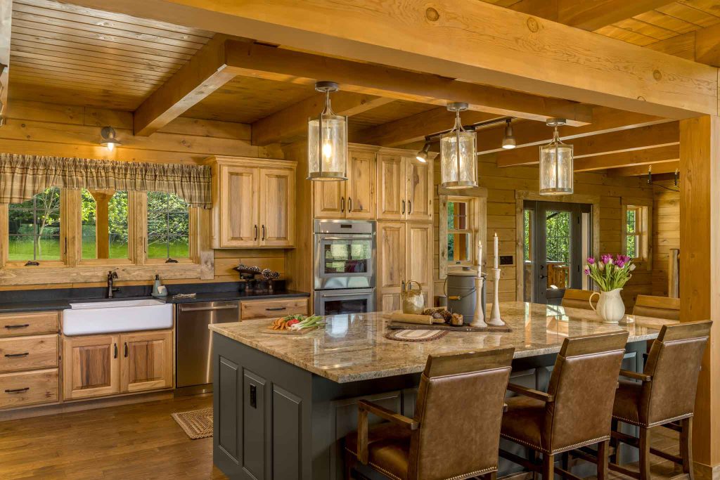 A spacious kitchen featuring wooden cabinetry, a large island with seating, and modern appliances. Sunlight streams through windows, illuminating lush greenery outside. Decorative pendant lights hang from the ceiling.