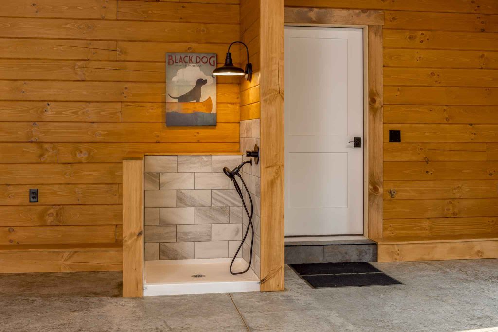 An outdoor shower area featuring a tiled base, a handheld showerhead, and a warm light fixture. The wooden walls are decorated with a poster of a black dog. A white door with a black handle is visible nearby.