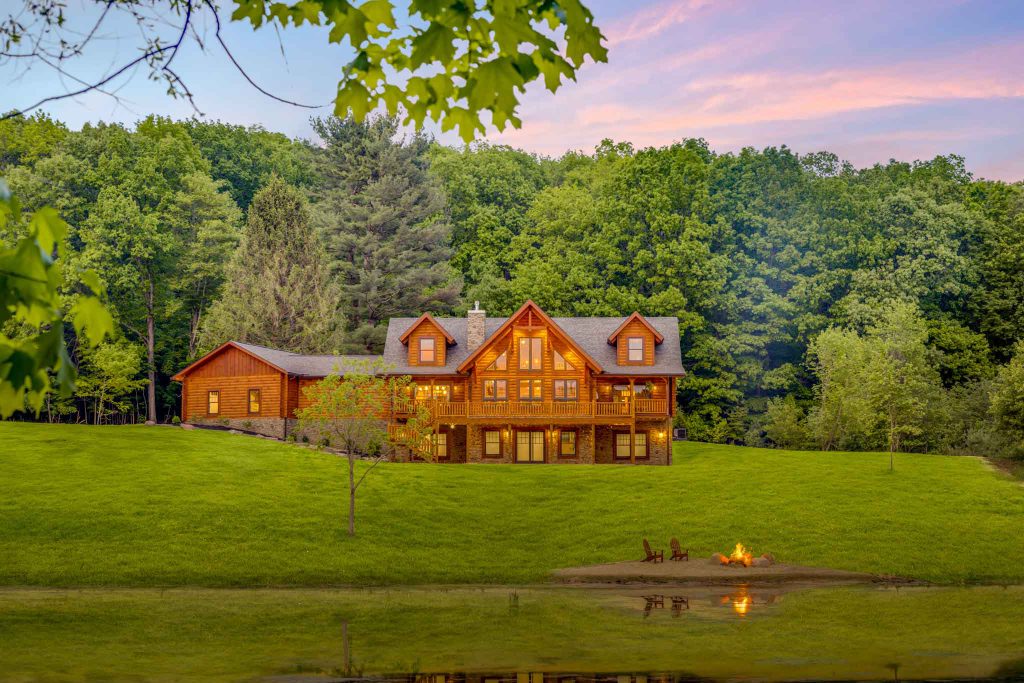 A rustic wooden cabin with large windows nestled beside a tranquil lake, surrounded by vibrant autumn foliage reflecting in the water. A fire pit with seating is visible on a sandy beach nearby.