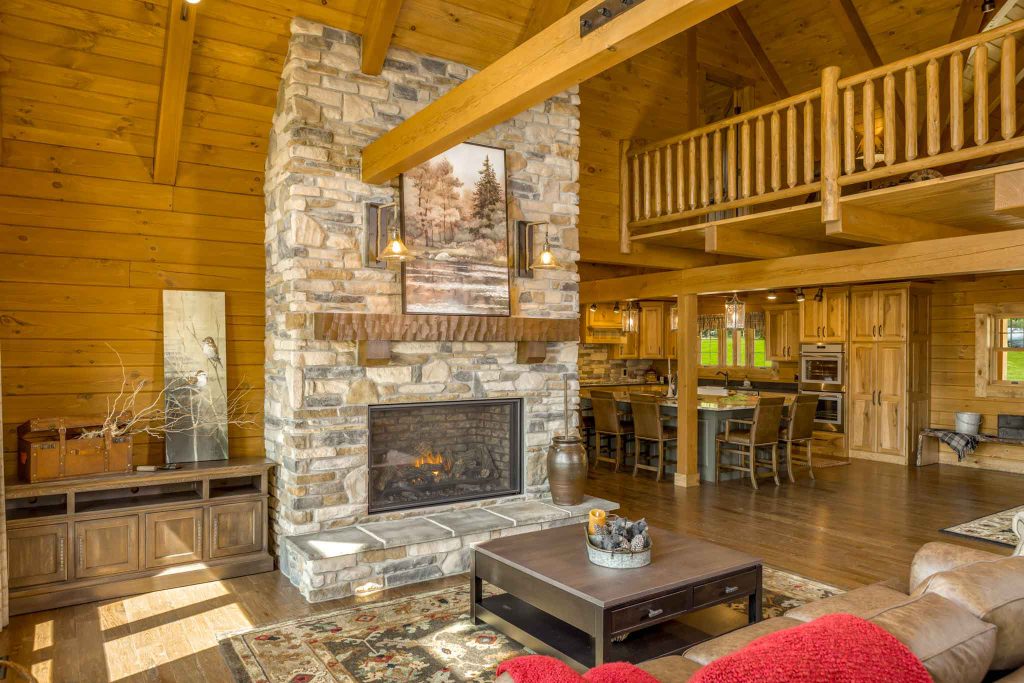 A cozy, rustic living room with wooden walls and a stone fireplace. The spacious area includes a brown sofa with a red throw, a wooden coffee table, and a dining area in the background, accented by warm lighting and a large window.