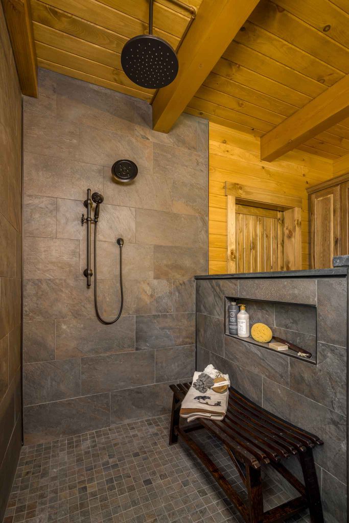 A modern shower space featuring a rainfall showerhead and a sleek, stone wall. A wooden bench sits beside a niche showcasing bath essentials, surrounded by warm wooden beams and a door leading outside.