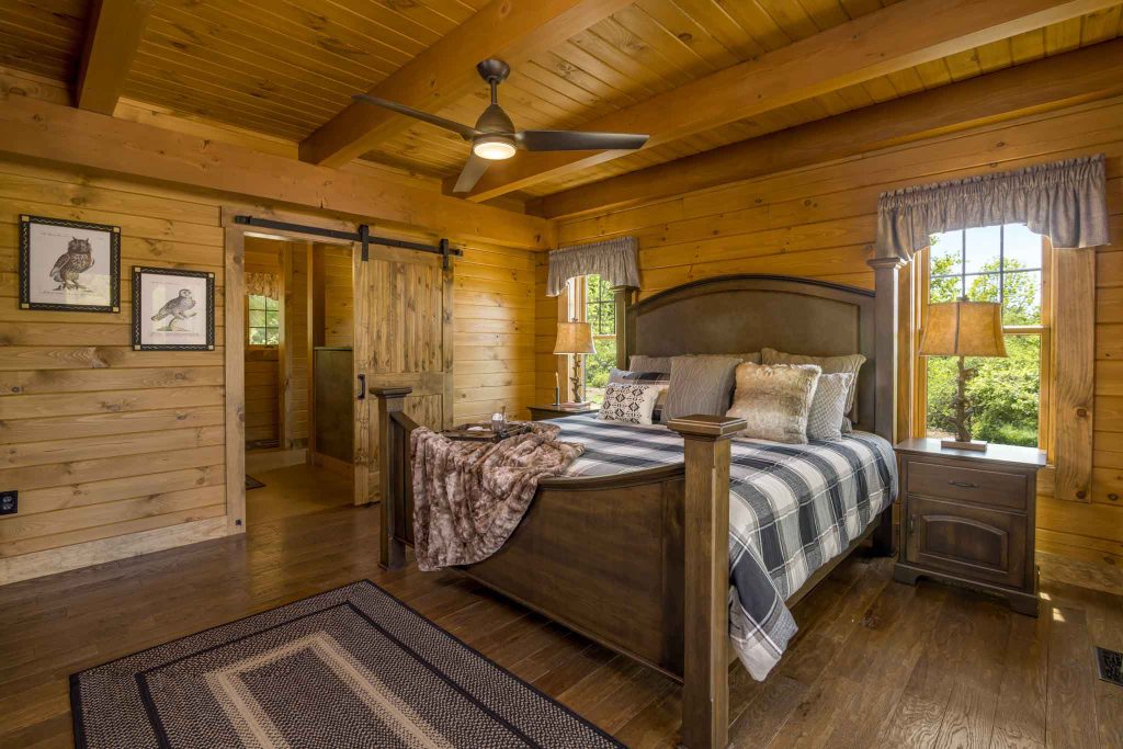 A cozy bedroom with wooden walls, featuring a large bed dressed in a plaid blanket, two bedside lamps on matching tables, and framed owl art on the walls. A visible sliding door leads to an adjoining space, and natural light shines through the windows.