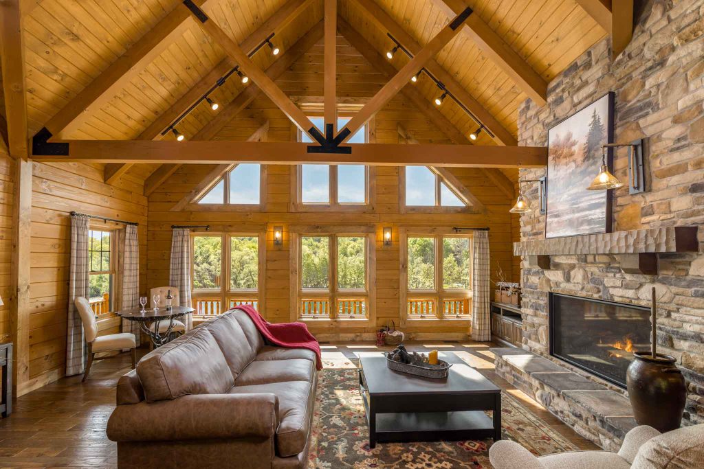 A cozy log cabin living room featuring a high wooden ceiling, stone fireplace, and large windows offering a view of greenery. A plush sofa with a red throw blanket sits near a wooden coffee table on a patterned rug.