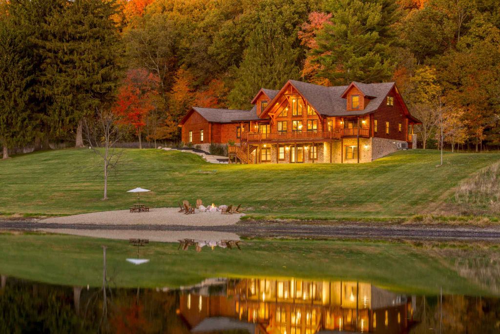 A rustic wooden cabin with large windows nestled beside a tranquil lake, surrounded by vibrant autumn foliage reflecting in the water. A fire pit with seating is visible on a sandy beach nearby.