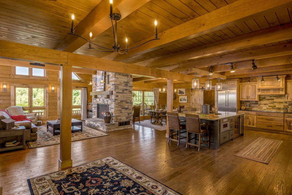 An inviting interior of a rustic cabin featuring wooden beams, a stone fireplace, and a spacious kitchen. The living area includes a cozy sofa and a dining table surrounded by wooden chairs, complemented by a decorative area rug.