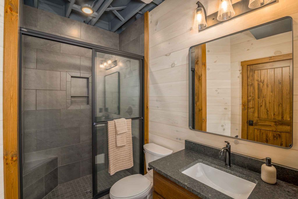 A modern bathroom featuring a glass shower enclosure, a white toilet, and a sleek vanity with a dark countertop. The space has wooden accents and a large mirror reflecting soft lighting, creating a welcoming atmosphere.