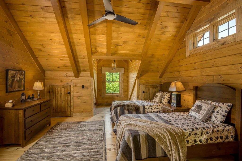 A cozy bedroom featuring two twin beds with patterned covers, wooden walls, and sloped ceilings. A rustic dresser and lamp are nearby, with a window providing natural light, enhancing the warm atmosphere of the space.