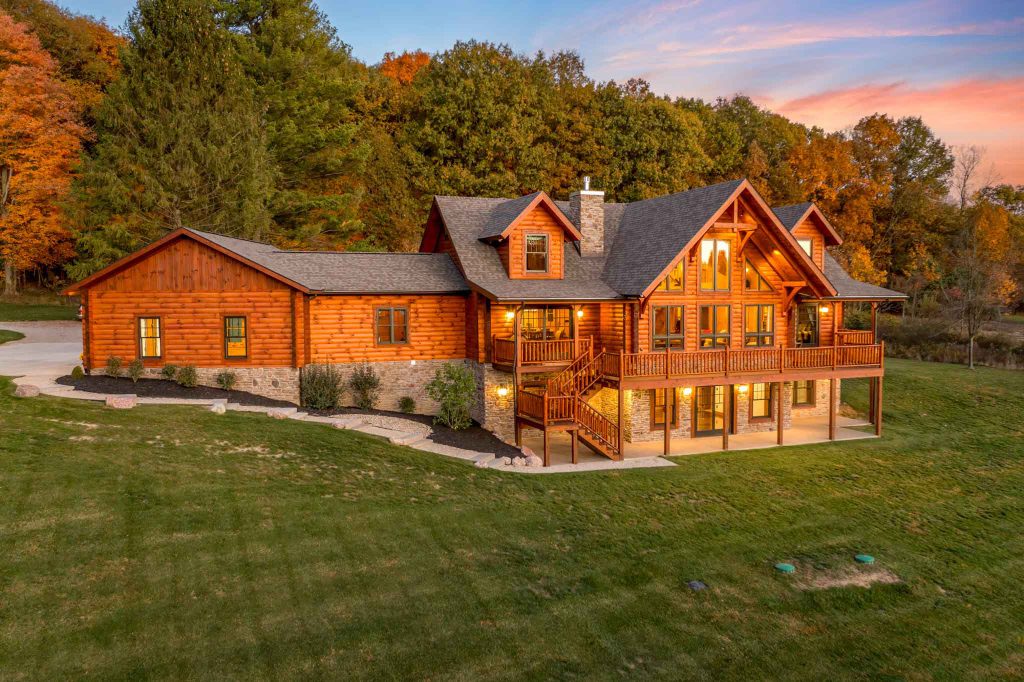 A large, elegant log cabin with a rustic wooden façade, set in a lush green landscape. The cabin features multiple balconies, large windows reflecting the autumn foliage, and a driveway leading to the entrance. The sky is painted in warm sunset hues.