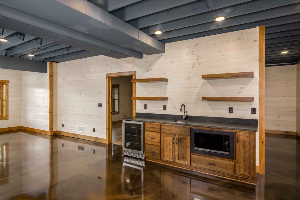 A spacious interior of a modern kitchen featuring wooden cabinets with a black countertop, a microwave, and open shelves. Polished concrete floors reflect the light, while exposed beams add to the contemporary rustic feel.