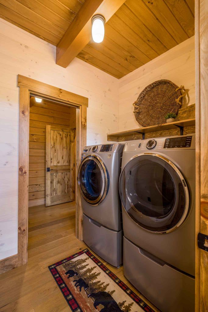 A cozy laundry room featuring two silver front-loading washing machines side by side, situated against a wooden wall. A decorative woven basket hangs above a shelf. A rustic patterned rug with bear motifs is laid on the wooden floor.