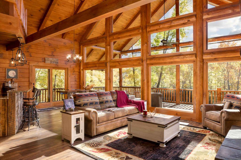 A cozy wooden cabin living room featuring large windows with wooden beams, a plush sofa with a blanket, a coffee table on a decorative rug, and a serene view of autumn trees outside.