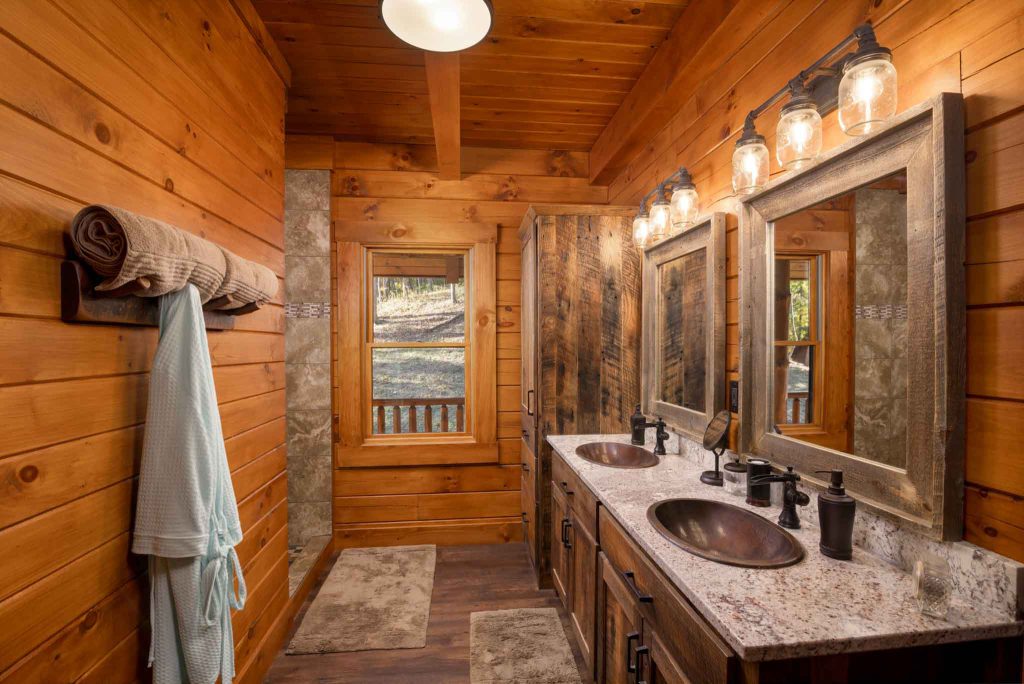 A cozy wooden bedroom with a king-sized bed accompanied by two bedside tables and lamps. A warm blanket drapes over the bed. Large glass doors open to a wooden deck, revealing a lush outdoor view.