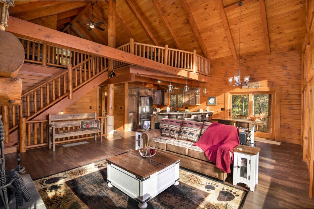 A cozy wooden cabin interior featuring a spacious living area with high ceilings, a comfortable sofa draped with a red blanket, a wooden coffee table, and a rustic staircase.