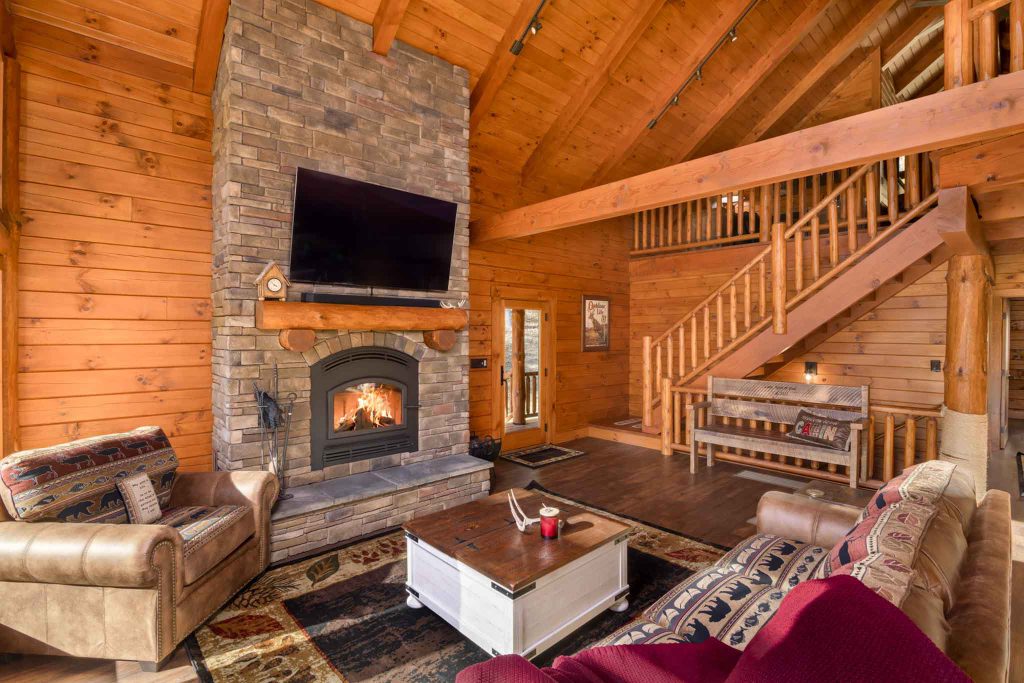 A cozy living room in a wooden cabin featuring a stone fireplace, a large flat-screen TV, and comfortable seating. A coffee table sits in the center, surrounded by rich rugs and wooden accents. Stairs lead to the upper level.