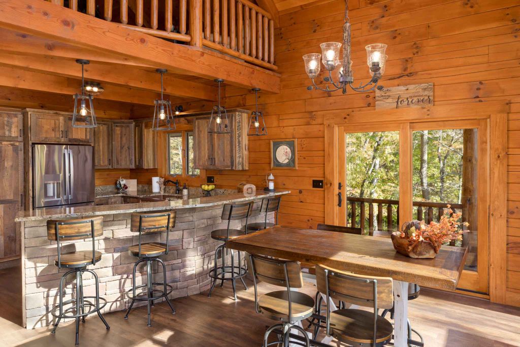 A cozy rustic kitchen featuring wooden cabinets and a granite countertop with bar stools. Sunlight floods in through large windows, illuminating a wooden dining table adorned with flowers. Scenic views of trees are visible outside.