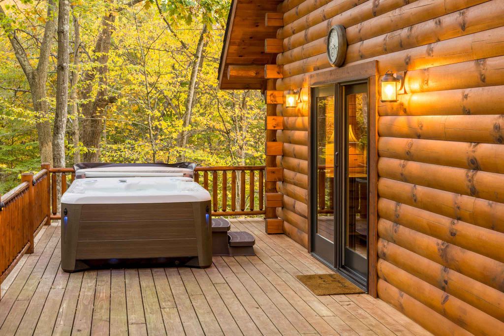 A wooden cabin deck surrounded by autumn foliage features a hot tub on the right side, with warm lighting illuminating the entrance. The rustic logs of the cabin contrast beautifully with the vibrant yellow leaves.