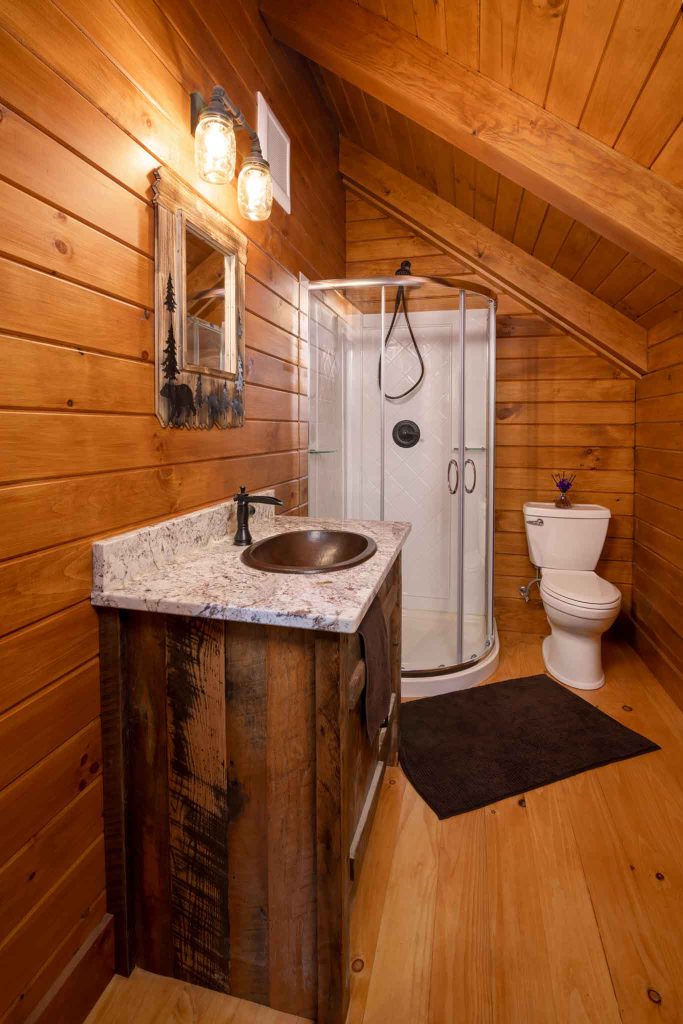 A cozy bathroom featuring wooden paneling, a rustic vanity with a marble countertop and a round sink, a glass shower enclosure, and a white toilet. The space is illuminated by vintage-style lighting. Light wooden flooring complements the warm atmosphere.