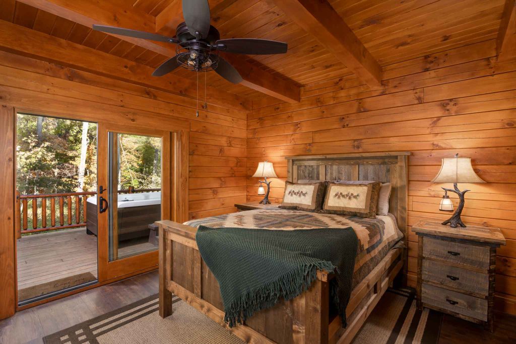 A cozy wooden bedroom with a king-sized bed accompanied by two bedside tables and lamps. A warm blanket drapes over the bed. Large glass doors open to a wooden deck, revealing a lush outdoor view.
