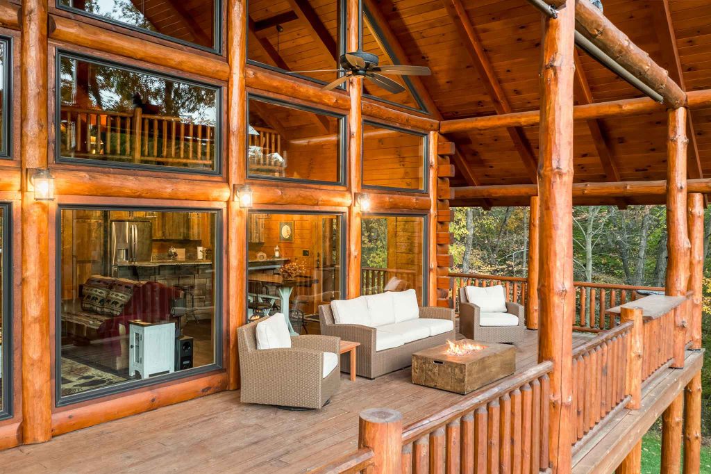 A spacious wooden deck of a log cabin featuring a cozy seating area with two armchairs and a sofa around a fire pit. Large windows reveal an inviting interior space, surrounded by trees and natural light.